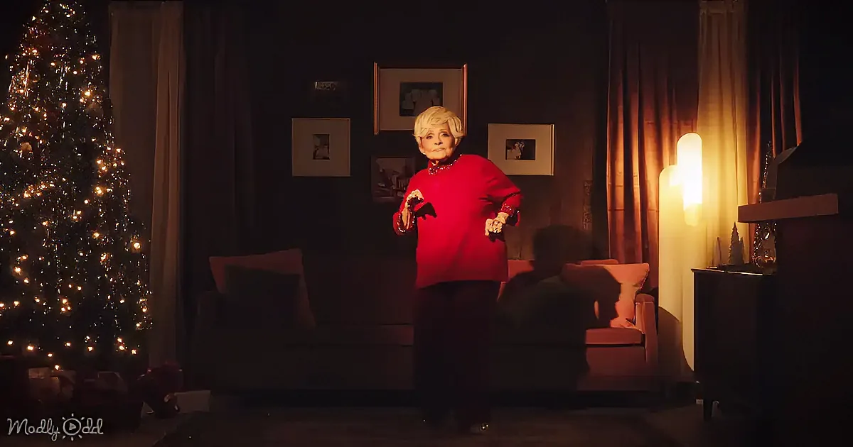 A woman in a red sweater stands in a dimly lit room with a Christmas tree, holding a small object.