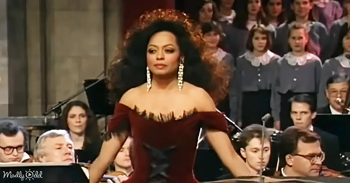 A woman in a red dress with a corset and large earrings stands in front of a choir and orchestra.