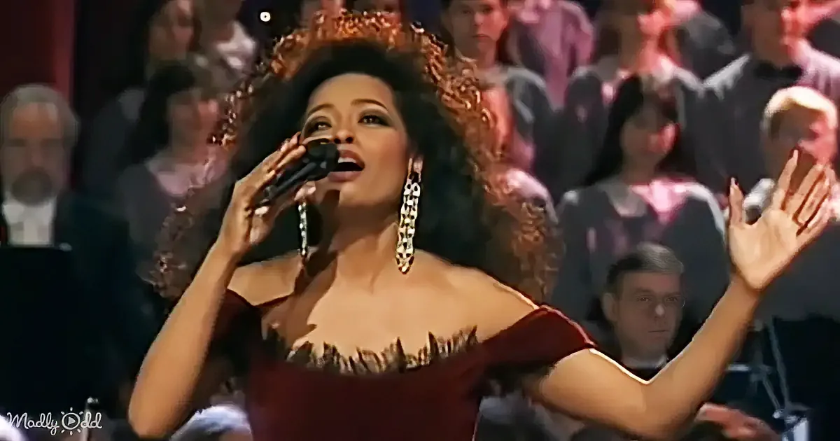 A woman in a red dress and ornate crown sings passionately into a microphone, surrounded by an audience.