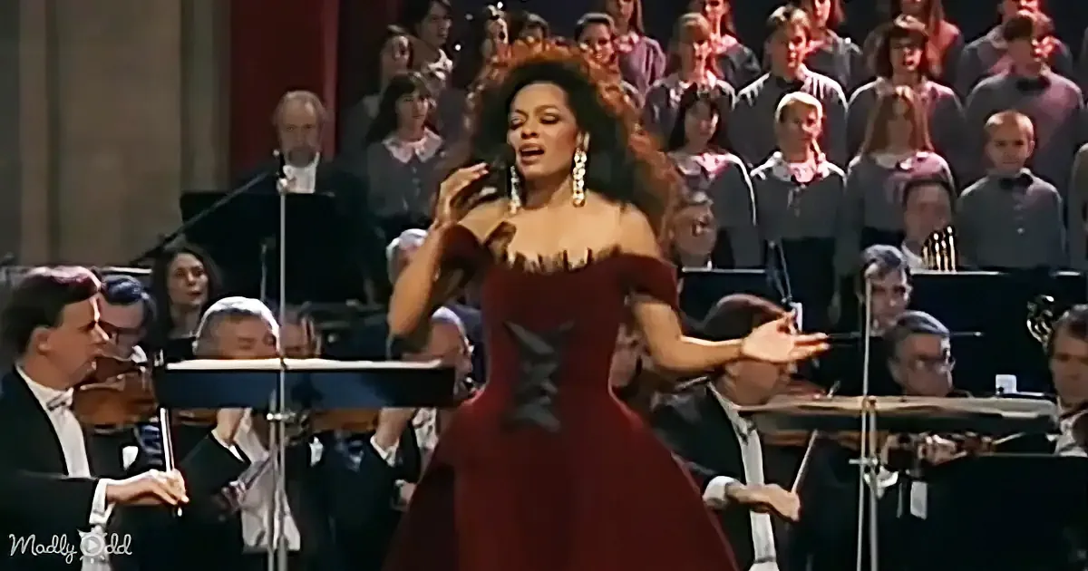 A woman in a red dress sings passionately on stage with an orchestra and choir in the background.