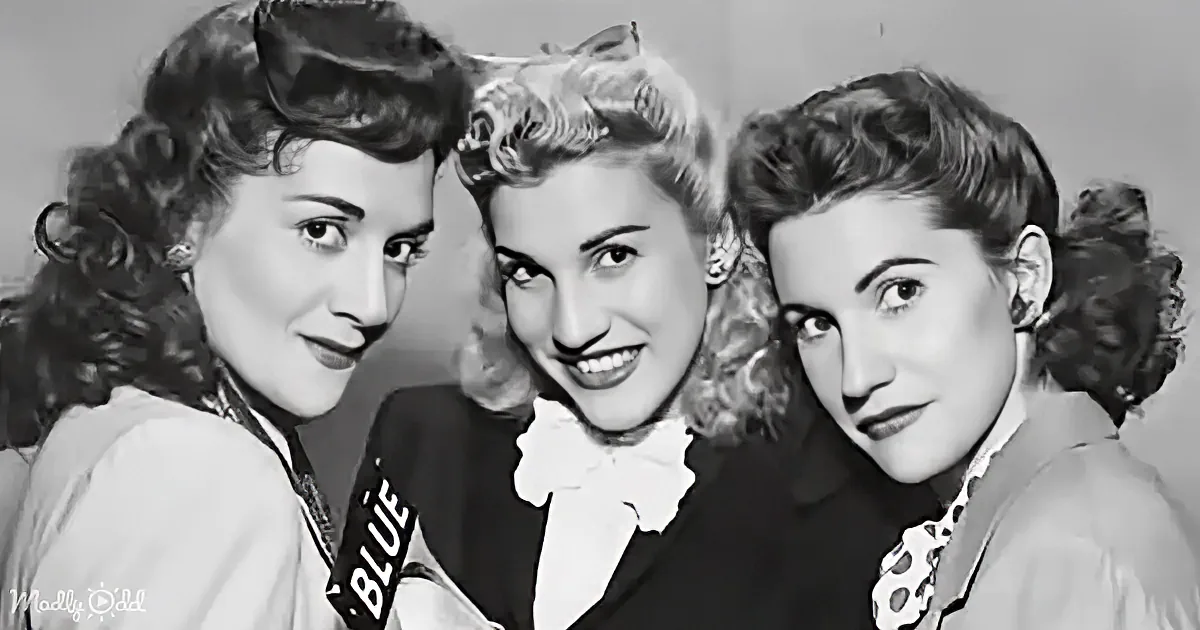 Three women with vintage hairstyles and clothing pose closely together, smiling.