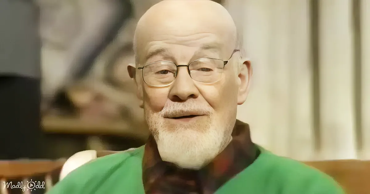 A bald man with glasses and a green shirt is smiling, sitting in a chair.