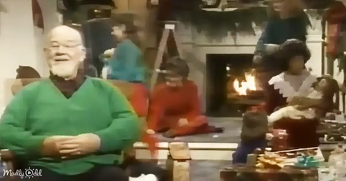 A man in a green sweater sits smiling, surrounded by others in a cozy room with a fireplace.