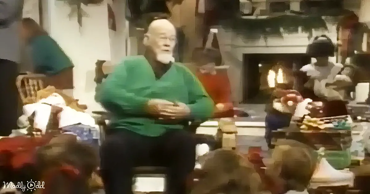 An older man in a green sweater sits in a chair, surrounded by people in a cozy room with a fireplace.