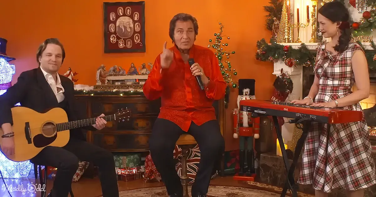 A man in red shirt sings into microphone, accompanied by guitarist and keyboardist in festive Christmas setting.