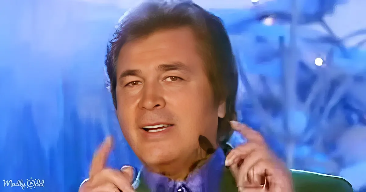A man with dark hair, wearing a blue shirt, is smiling and gesturing with his hands.