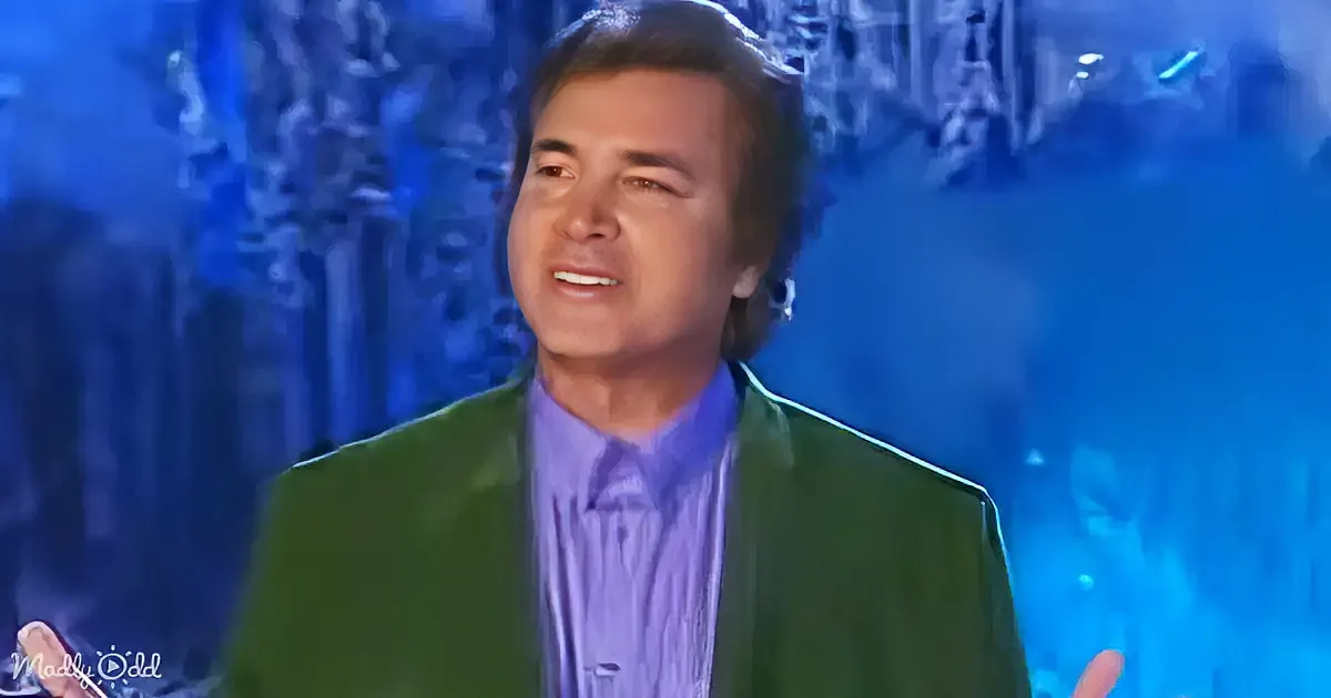 A man in a green suit and purple shirt stands against a blue-lit background, smiling.