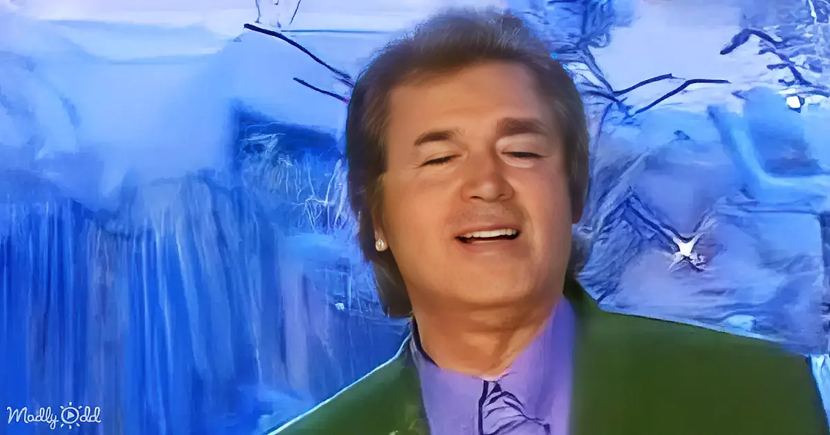 A smiling man in a green suit and purple shirt stands against a blue, abstract background.