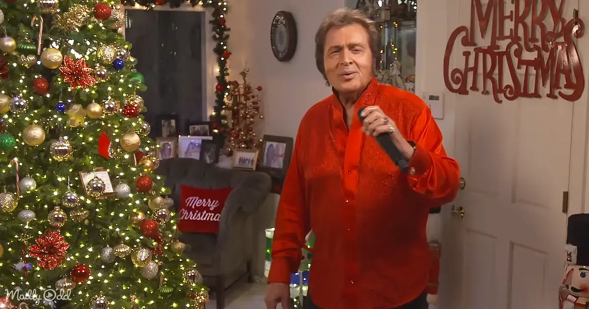 A man in a red shirt sings into a microphone near a decorated Christmas tree.