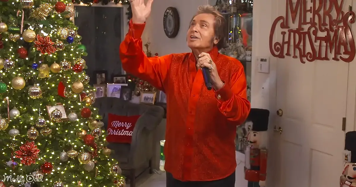 A person in a red shirt sings into a microphone, gesturing near a decorated Christmas tree.