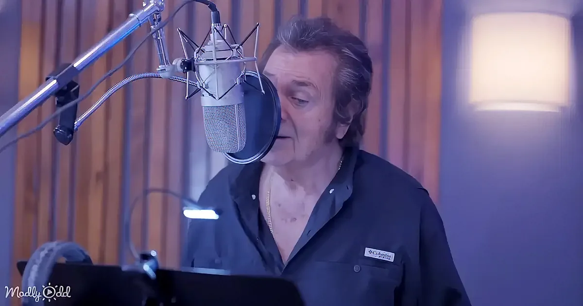 A man in a recording studio sings into a microphone, wearing a dark shirt and a gold chain.