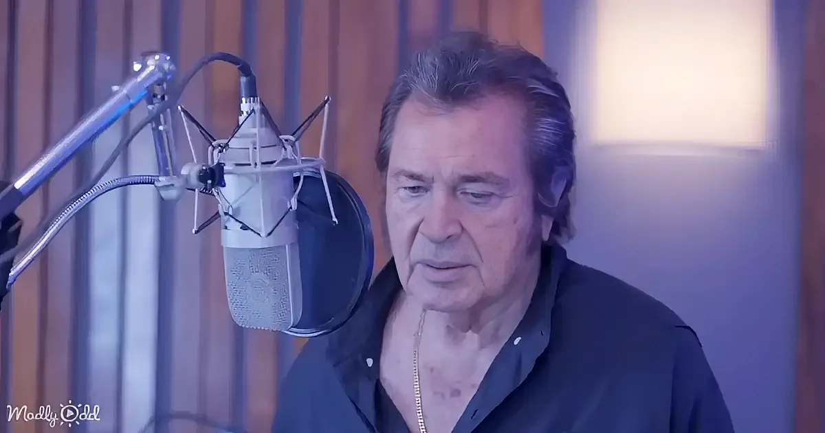 A man in a recording studio, wearing a black shirt, sits in front of a microphone, appearing focused.