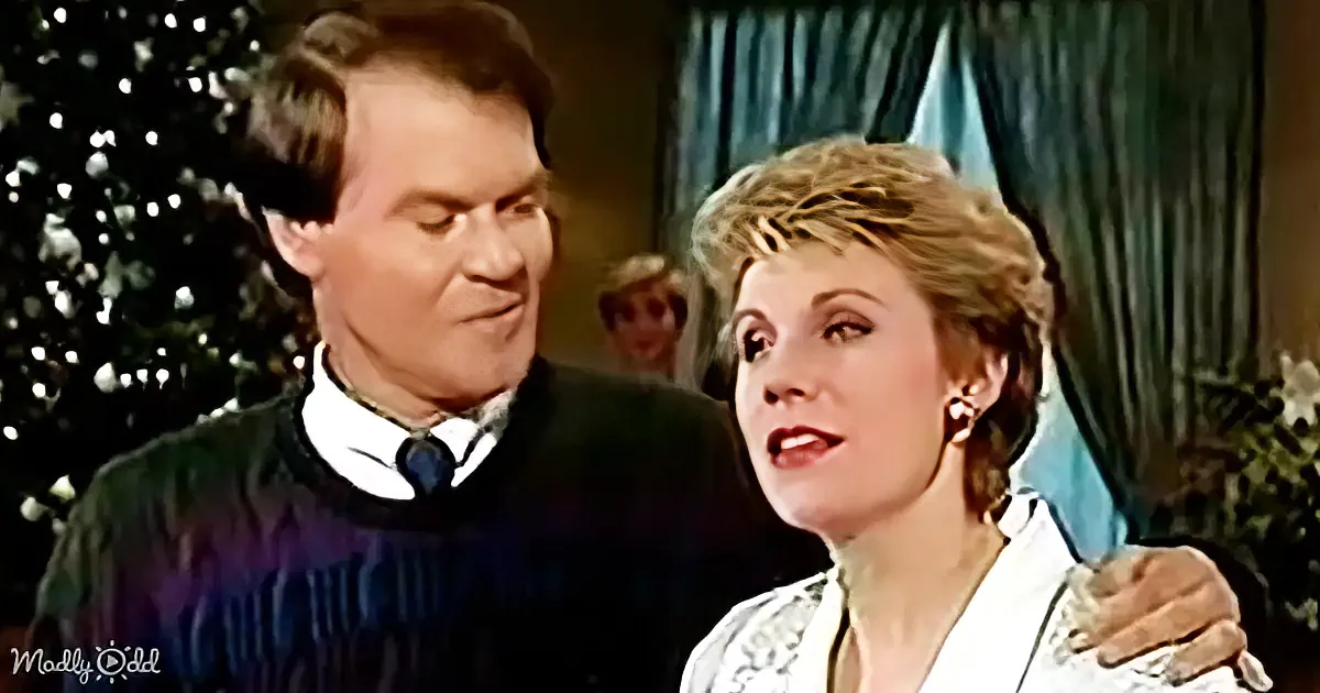 A man and woman are standing close, the man in a dark sweater, the woman in a light blouse, both looking at each other.