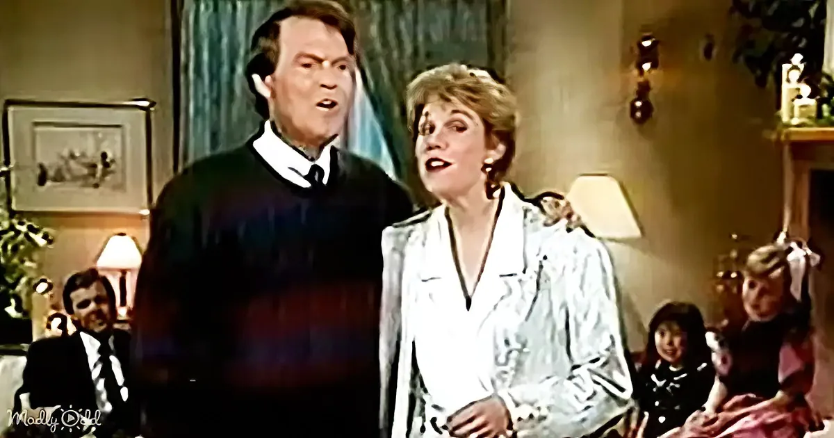 A man and woman are standing together, seemingly surprised or shocked, in a warmly lit living room.