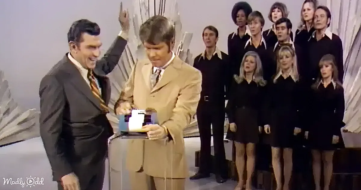 A man in a tan suit holds a cake while another man in a dark suit raises his hand, with a group of people in the background.