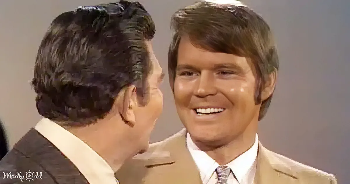 A man in a tan suit and white shirt smiles while another man in a dark suit looks at him.