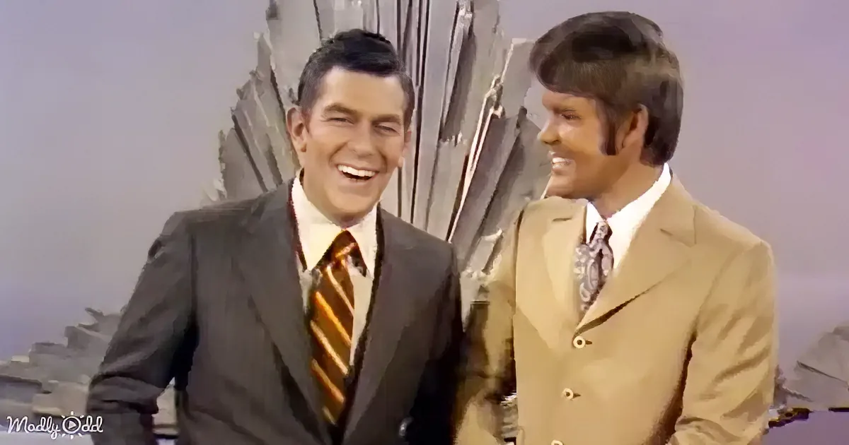 Two men in suits smile and laugh, one in a dark suit with a striped tie, the other in a beige suit with a patterned tie.