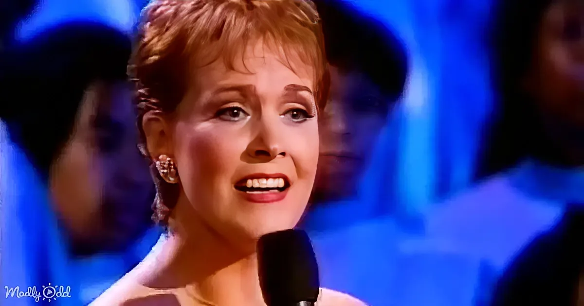 A woman with short red hair and earrings sings into a microphone, smiling with a blue-lit background.