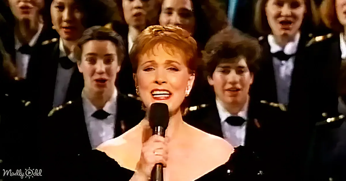 A woman in a white dress sings into a microphone, surrounded by a choir in formal attire.