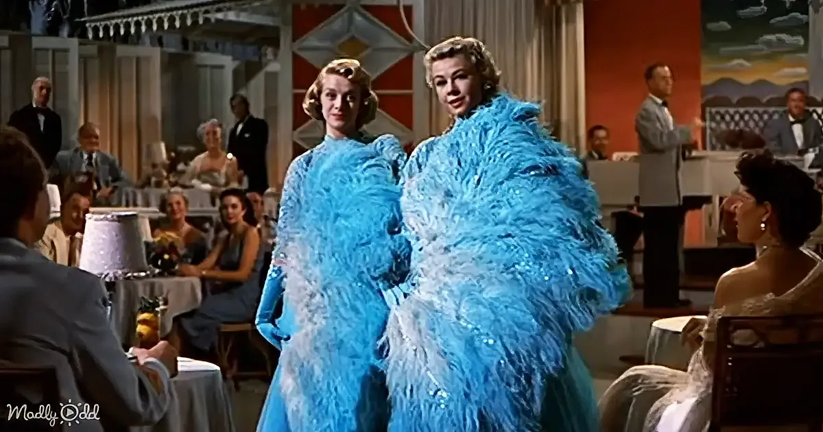 Two women in elaborate blue feathered gowns walk through a bustling restaurant, drawing attention.