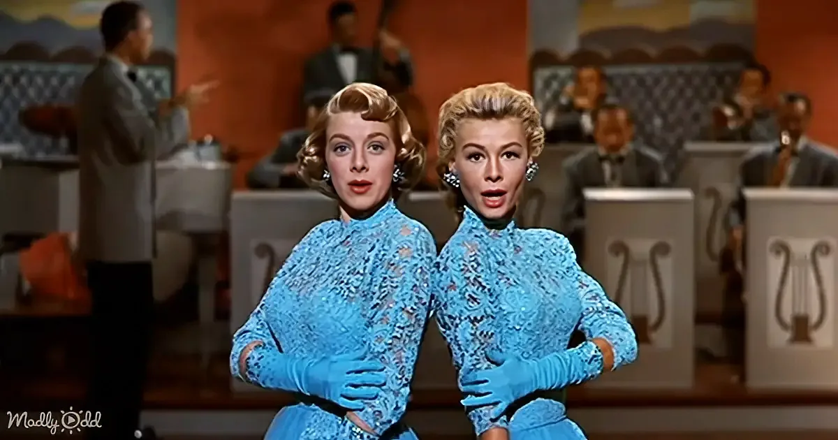 Two women in matching blue outfits with gloves, standing in front of a band, with a "Madly Odd" watermark.