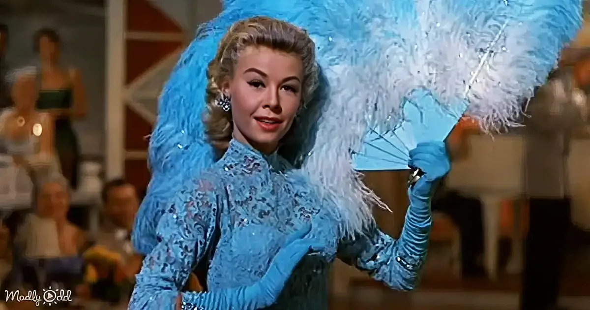 A woman in a blue sequined outfit holds a feathered fan, standing in a performance setting with blurred audience in the...