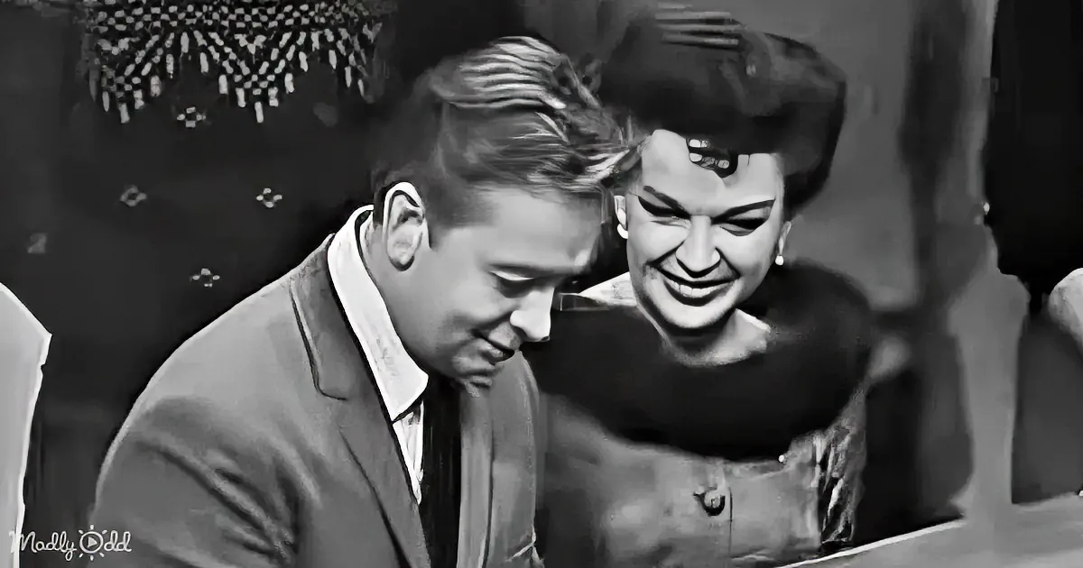A man in a suit leans forward, smiling, while a woman in a fur coat and hat looks on, both in a vintage setting.