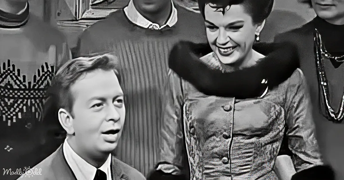 A man in a suit and tie smiles, while a woman in a patterned dress and fur collar looks down, surrounded by others.