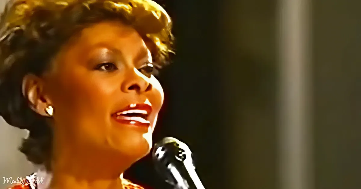 A woman with curly hair and earrings sings into a microphone, her face illuminated.