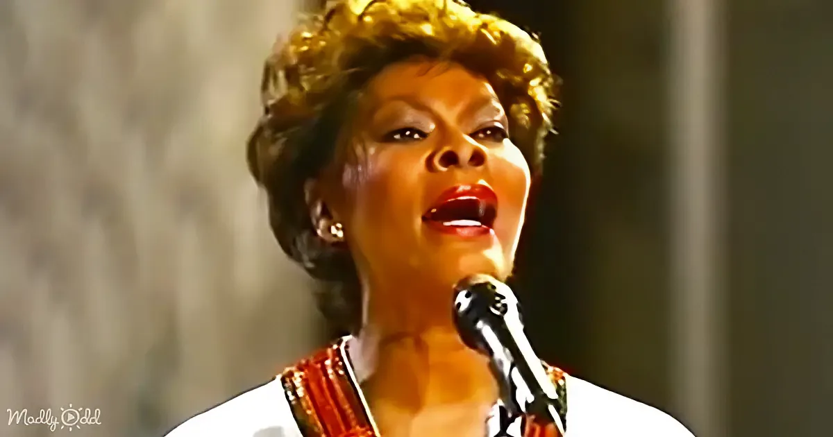 A woman with curly hair sings into a microphone, wearing a white top with red accents.