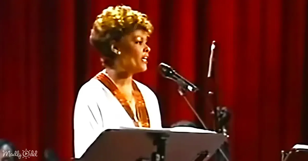 A woman with short, curly hair sings into a microphone on stage, wearing a white top and a gold necklace.