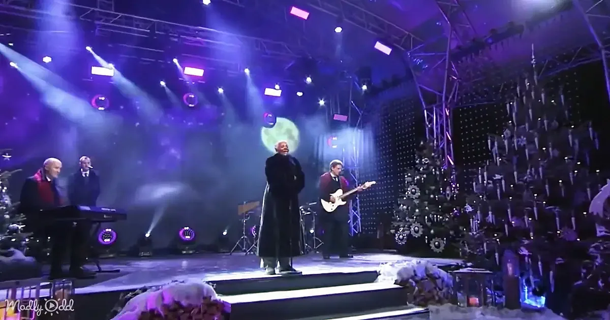 A man in a long coat stands on stage with musicians, surrounded by Christmas trees and purple lights.