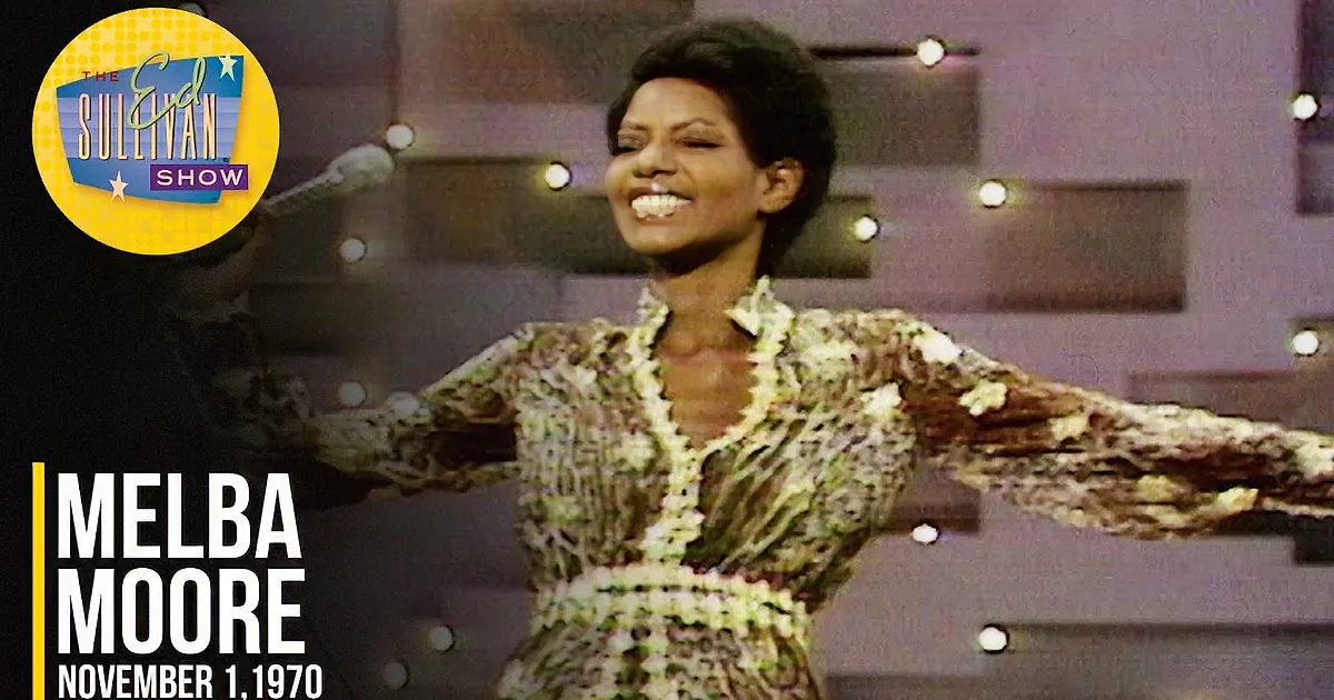 A smiling woman with short hair, wearing a patterned top, holding a microphone, with blurred lights in the background.