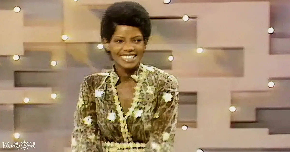A woman in a floral dress smiles on a stage with a geometric background and lights.