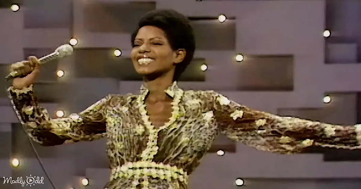 A woman in a patterned dress holds a microphone, smiling, with outstretched arms, against a backdrop of lights and a dark...