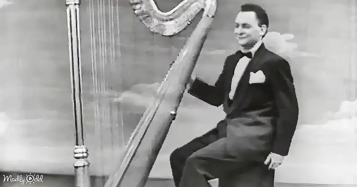 A man in a tuxedo sits beside a harp, smiling, in a black-and-white image.
