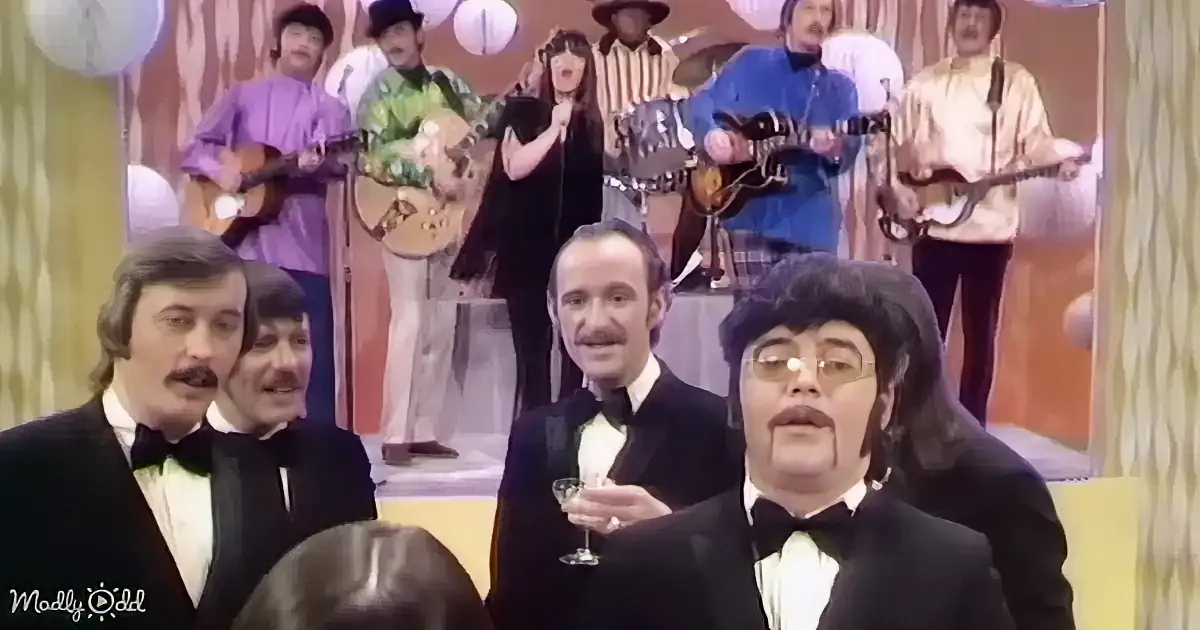 A man in a tuxedo holds a glass, surrounded by other formally dressed individuals and a band playing in the background.