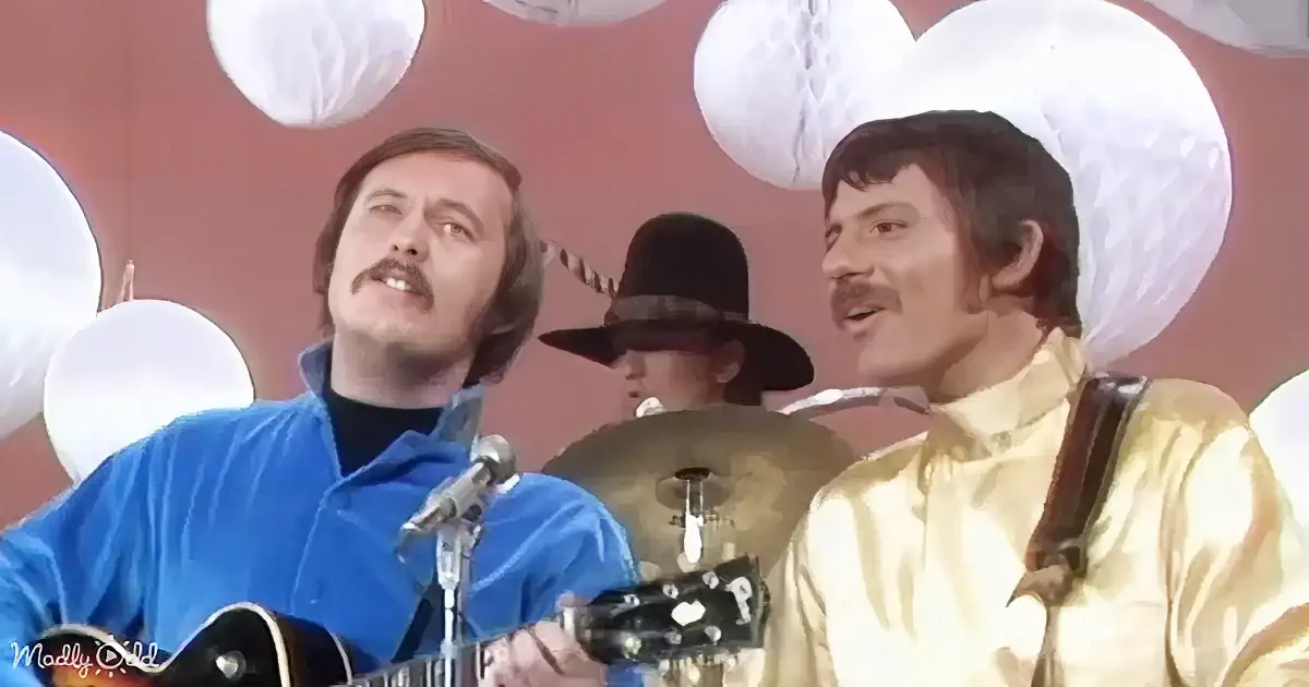 Two men in vibrant outfits play music on stage with a drummer in a hat behind them.