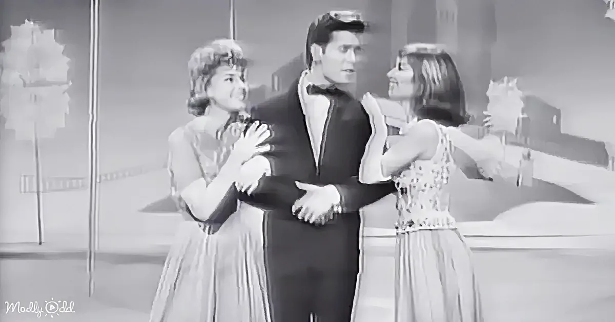 A man in a tuxedo is embraced by two women in dresses, set against a cityscape backdrop.
