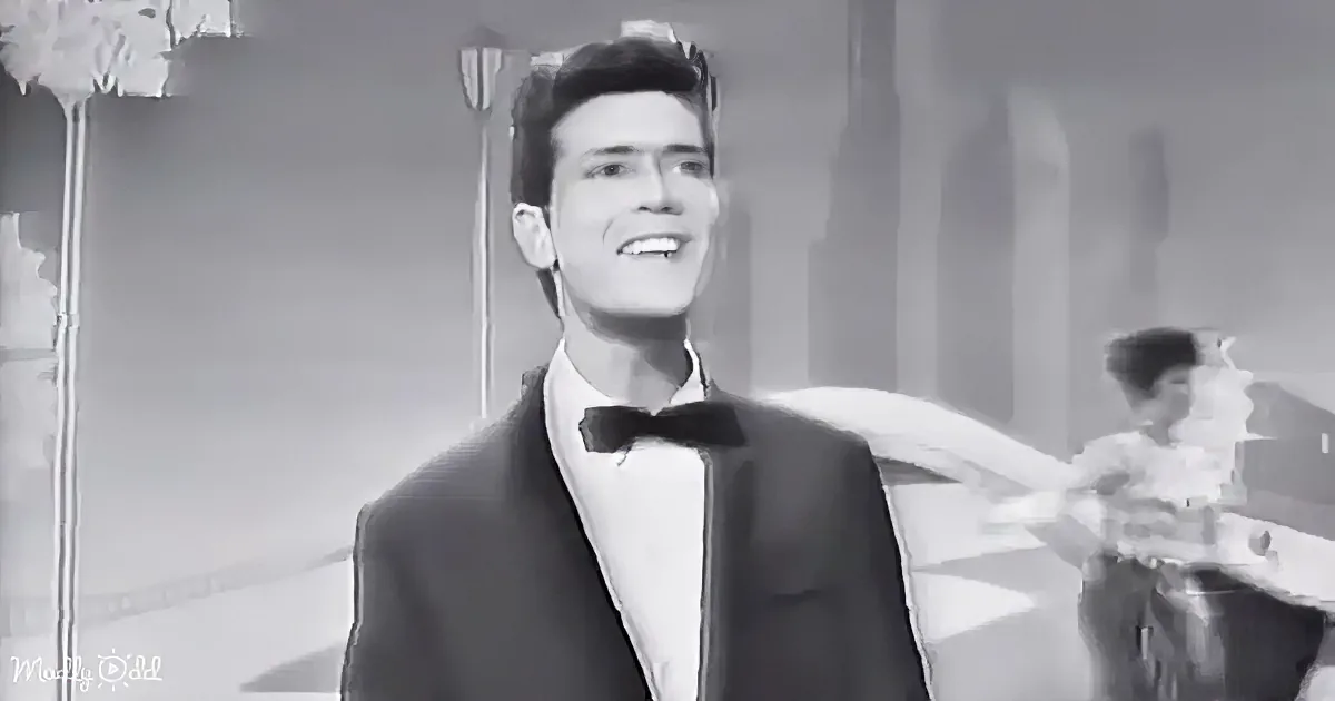 A man in a tuxedo smiles, standing in a black-and-white scene with a dancer in the background.
