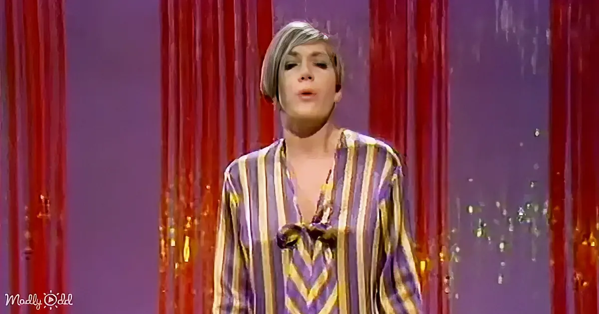 A person in a striped shirt stands on stage with red curtains and purple background.