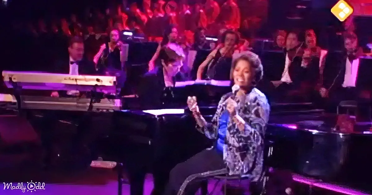A woman in a patterned dress sings into a microphone on stage, surrounded by musicians and a vibrant audience.