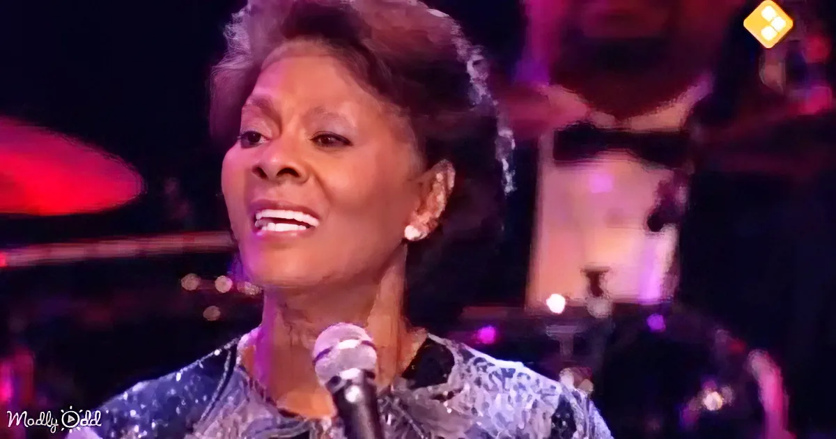 A woman with short, curly hair smiles while singing into a microphone on stage.