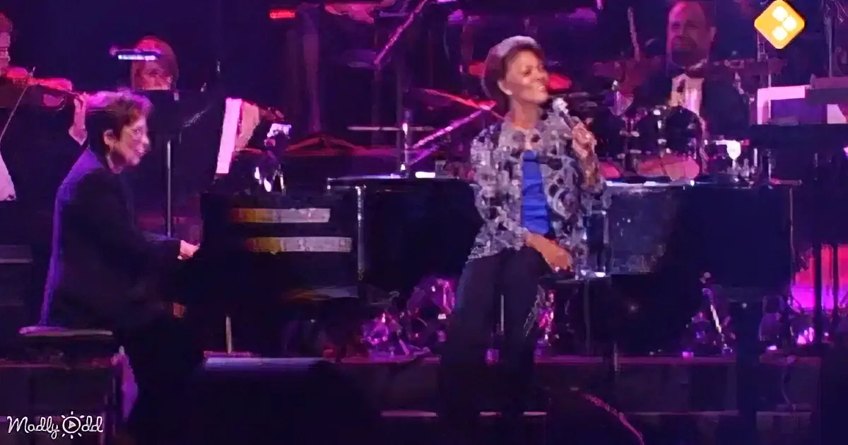 A woman sings passionately into a microphone on stage, accompanied by a pianist and a band.