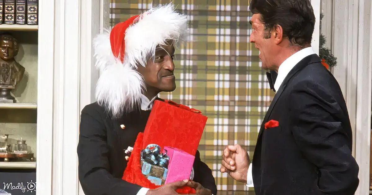A man in a Santa hat holds a gift, talking to another man in a suit, both in a festive setting.