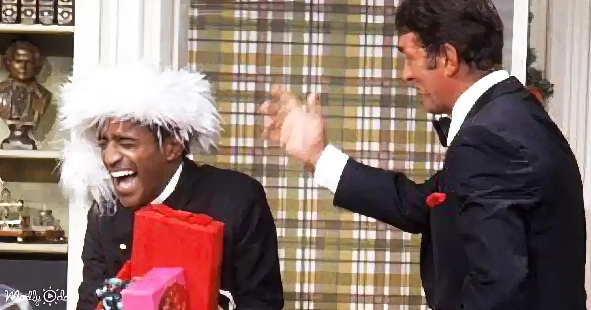 A man in a white fur hat and suit holds a red gift, laughing, as another man in a suit gestures excitedly.