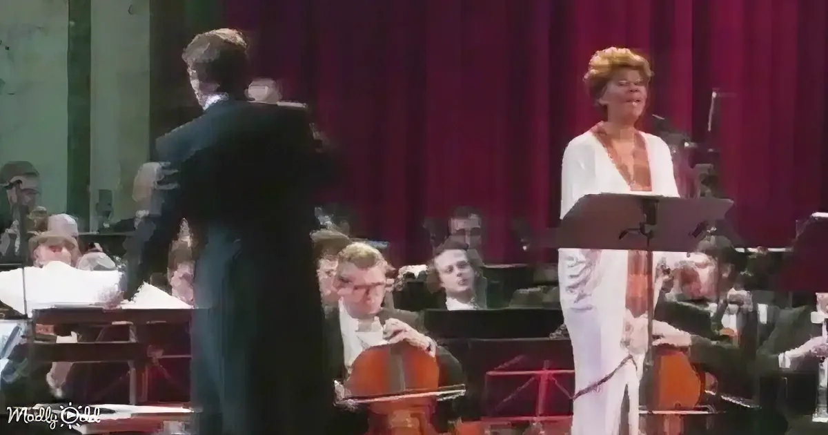 A conductor leads an orchestra with a female singer in a white dress at a podium.