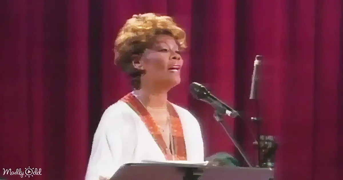 A woman with short, curly blonde hair sings into a microphone on stage, wearing a white and orange outfit.