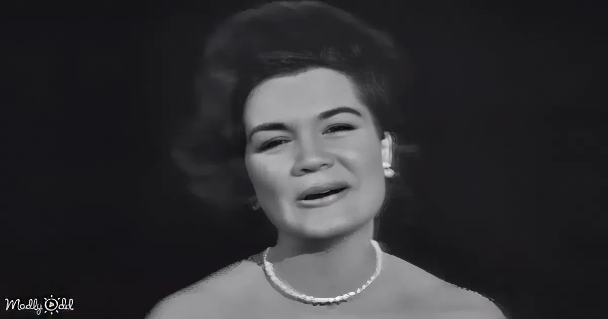 A smiling woman in a vintage outfit with a pearl necklace and earrings, holding her hands together.