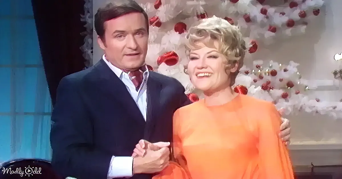 A man in a suit and a woman in an orange dress are smiling and holding hands in front of a Christmas tree.